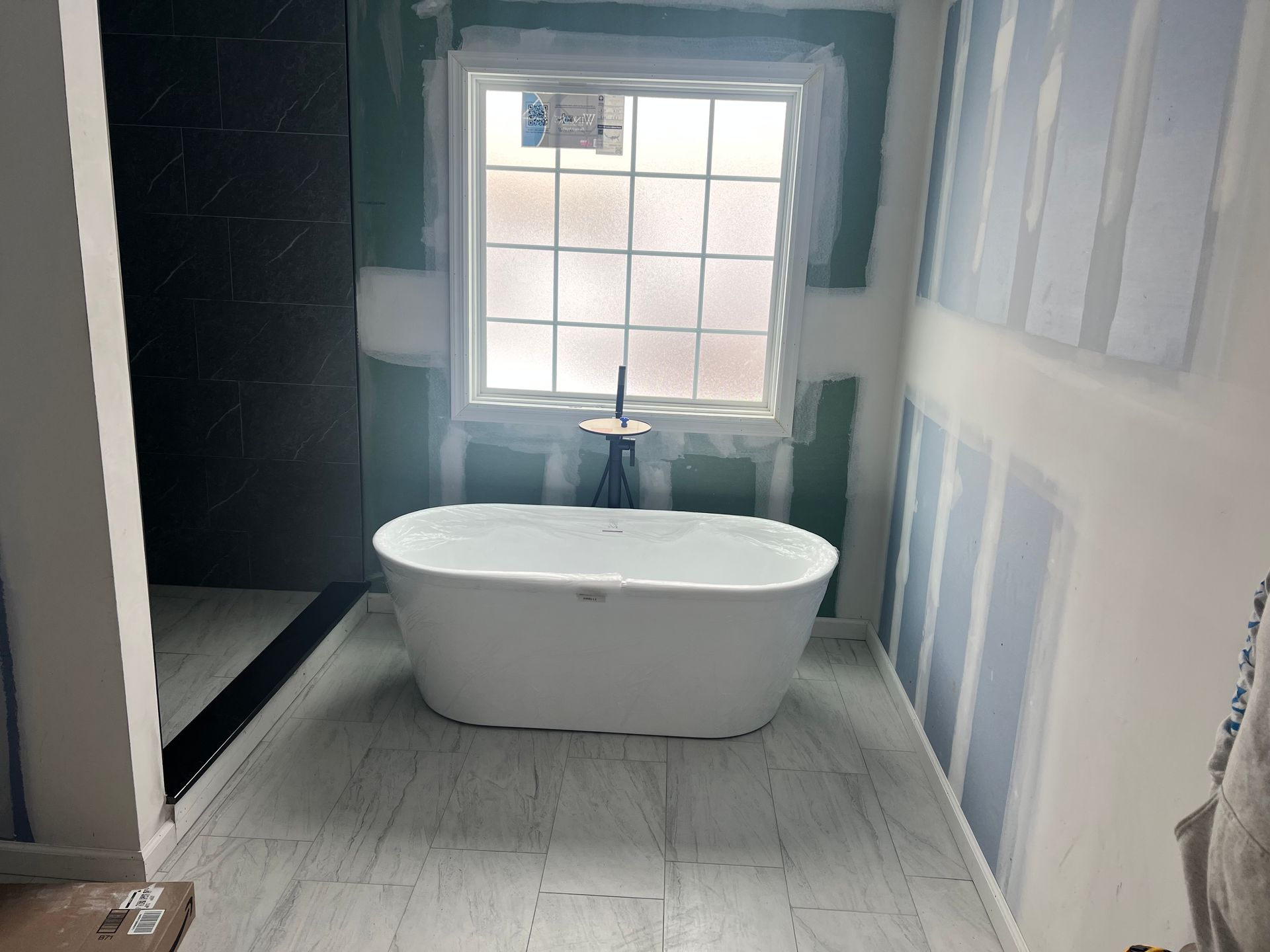 Bathroom under construction with a white bathtub in front of a window. Drywall and tiles are visible.