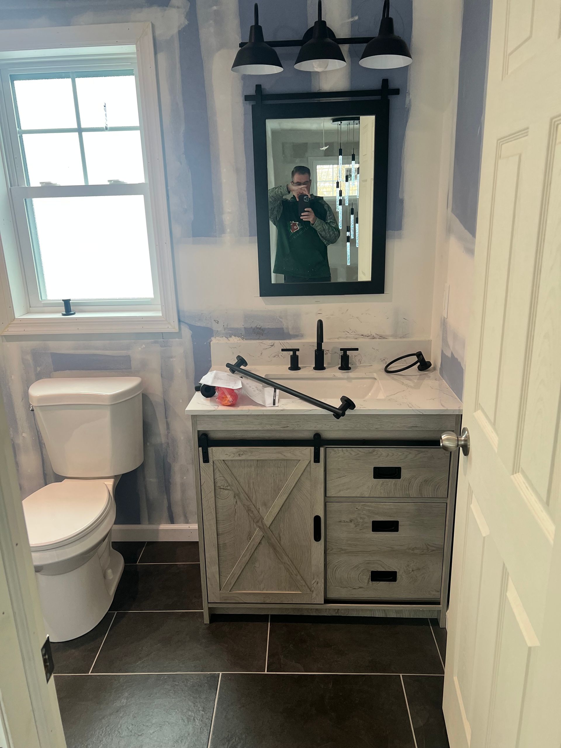 Bathroom with white toilet, vanity, black mirror, lights, and a person taking a photo. Blue walls, brown floor.