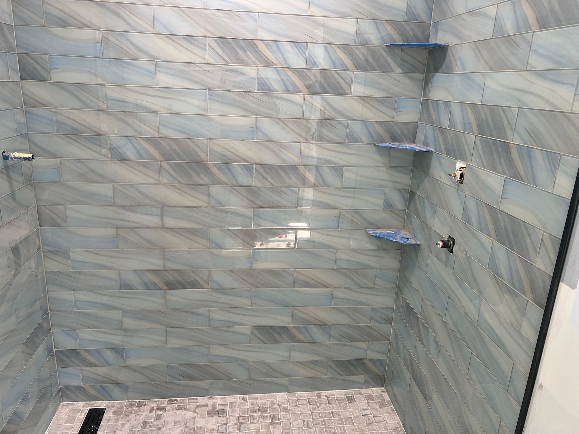 Blue tiled shower with wave-like pattern, two shelves, and fixtures.