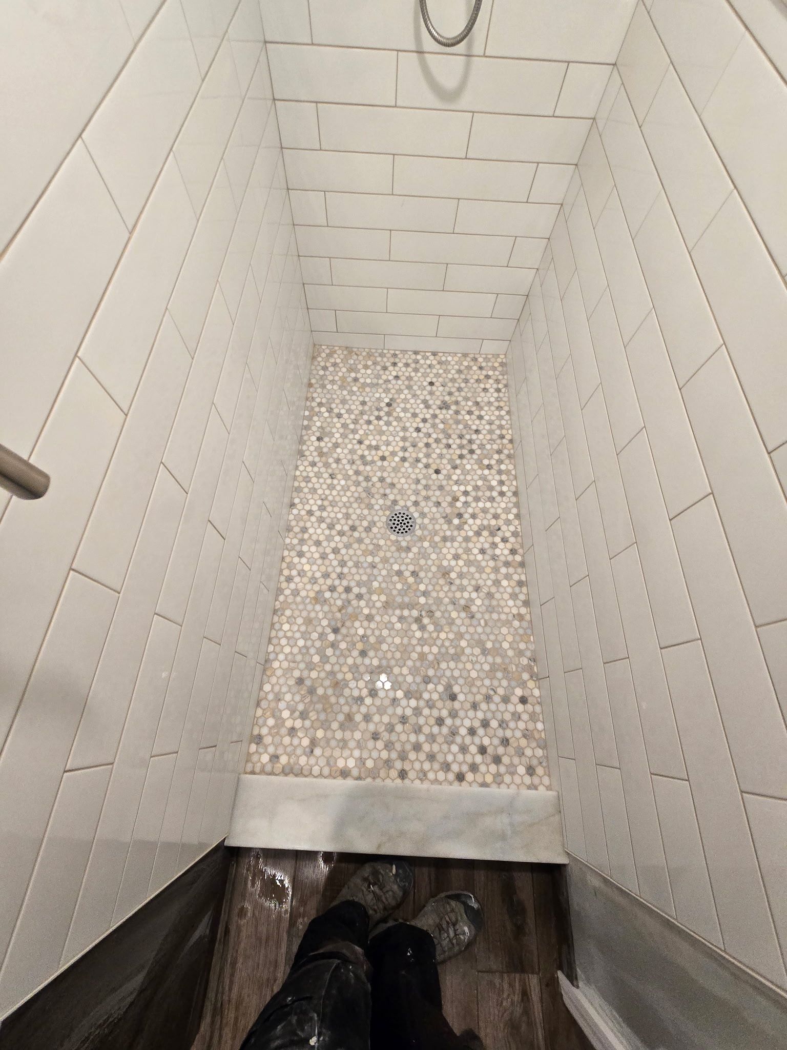 Narrow shower with white tiled walls, mosaic floor, and a person's feet in view.