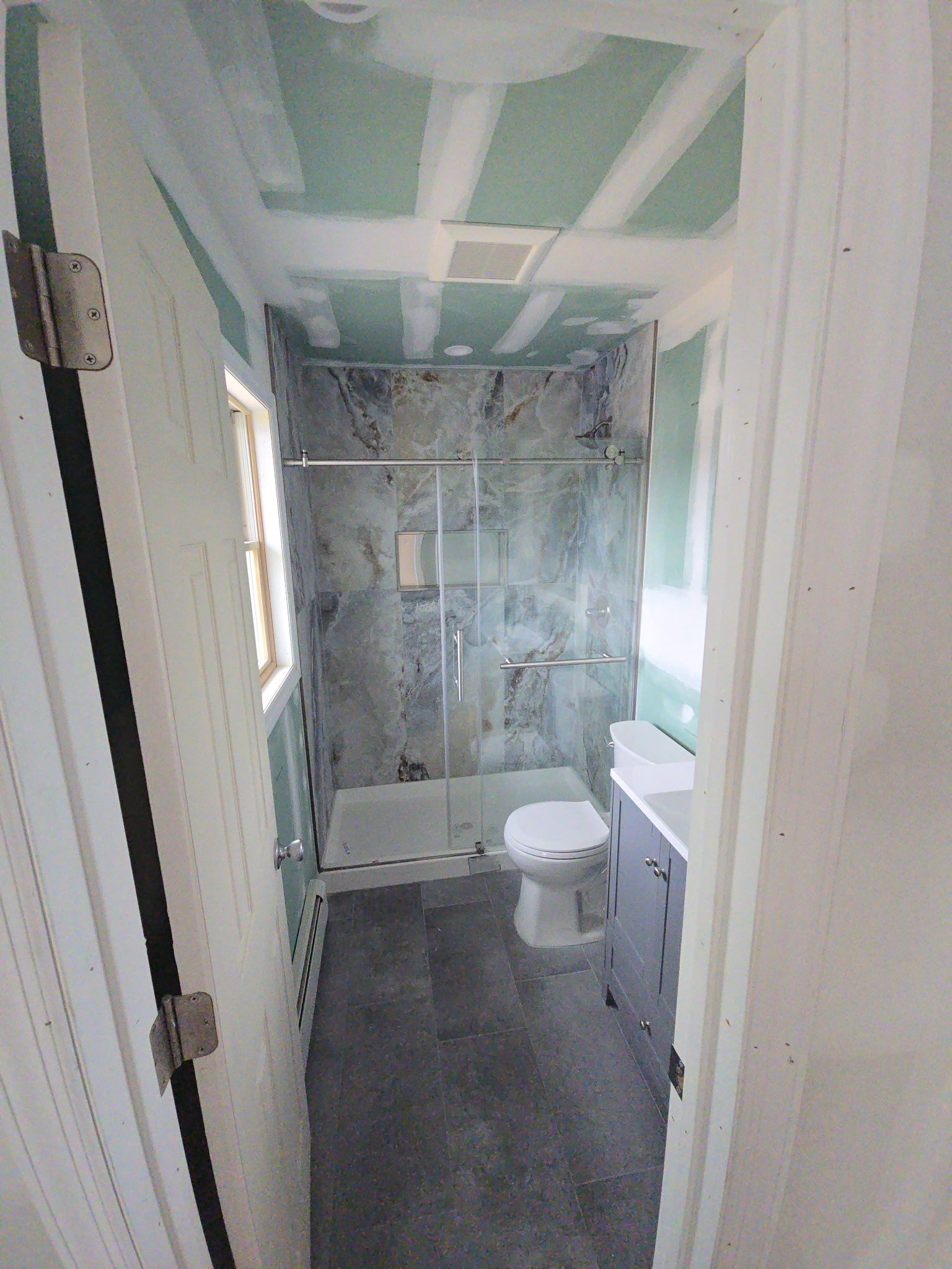 Bathroom under construction; shower, toilet, and vanity installed. Walls and ceiling have exposed drywall.