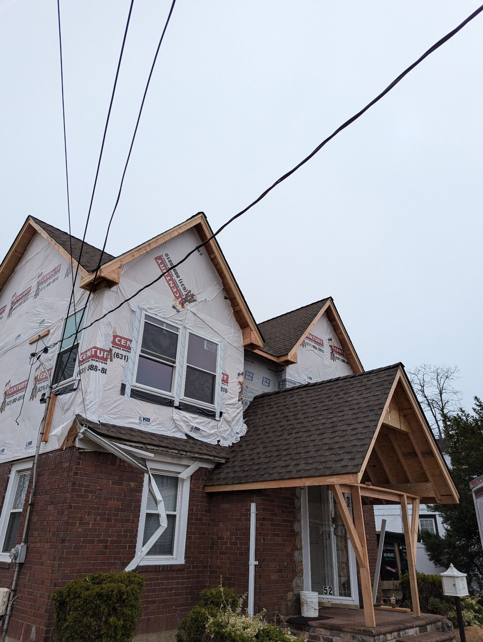 House under construction, covered with white wrap, brick facade, and exposed wooden roof frames. Power lines overhead.