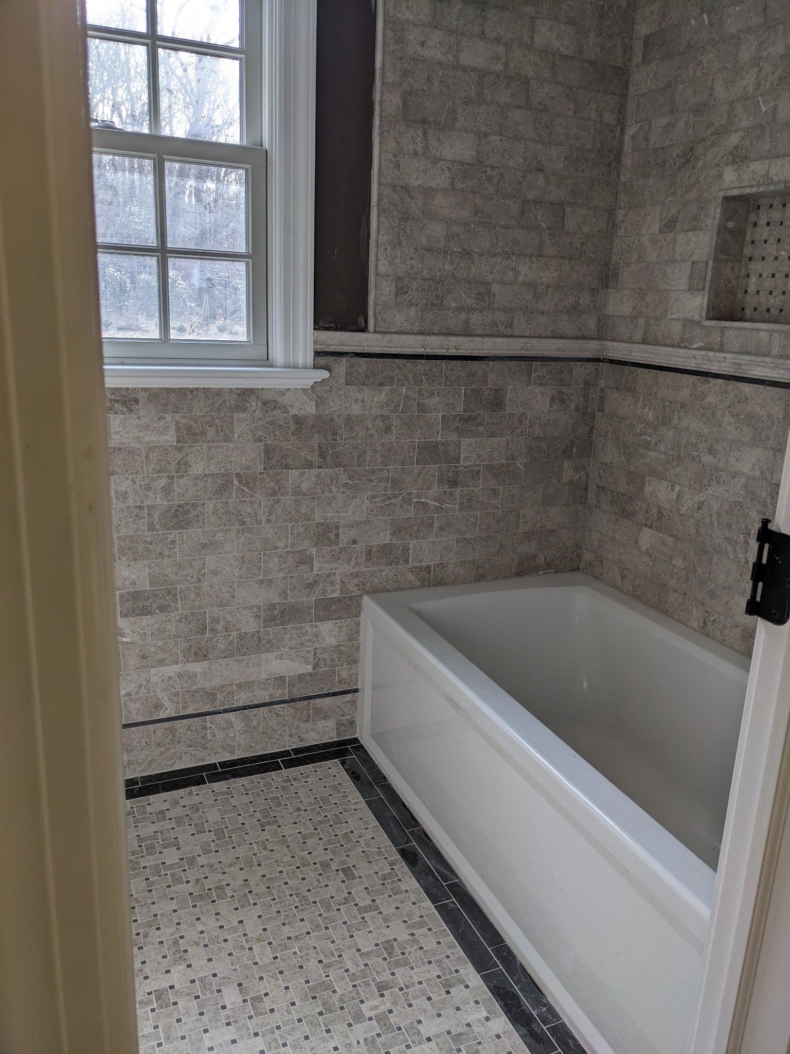 Bathroom with gray brick-like tile, white bathtub, window, and patterned tile floor.