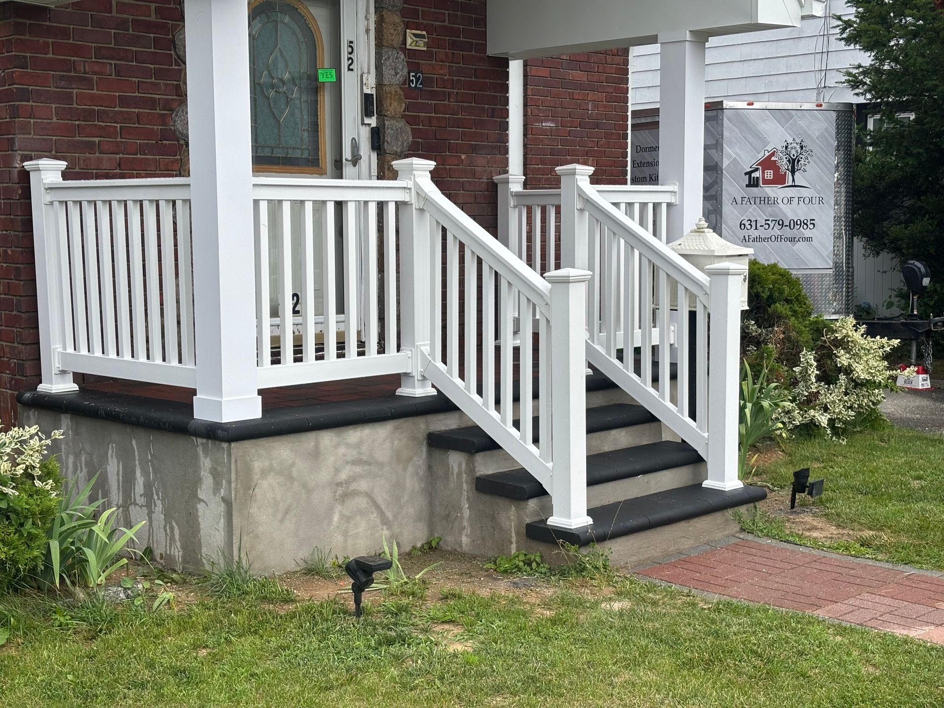 White railing and steps leading up to a brick building's porch with green grass and a red brick walkway.