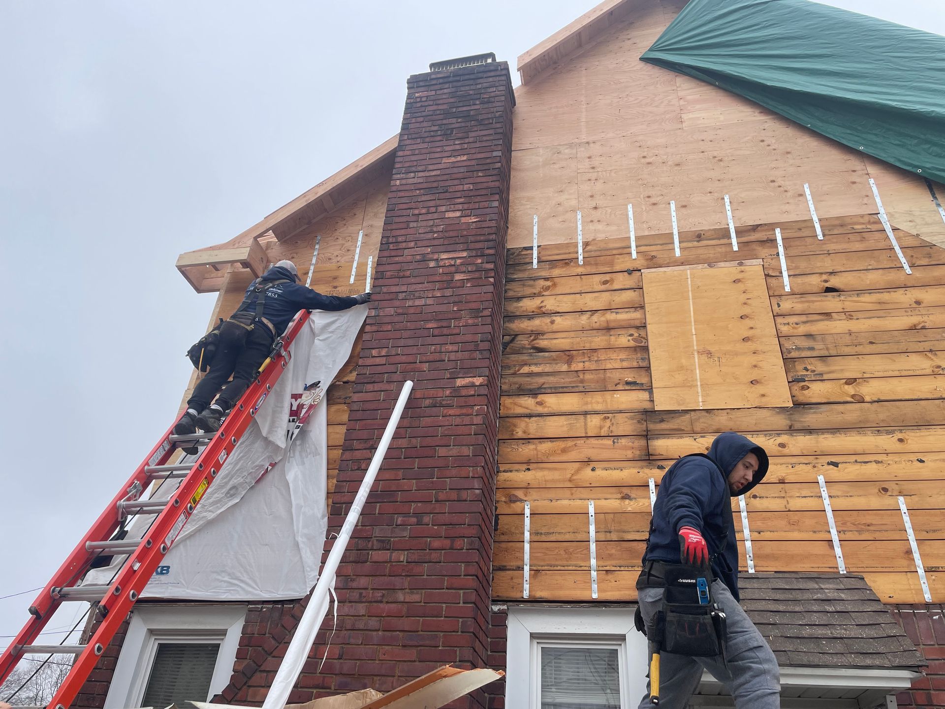 Two workers on a house. One on a ladder near the chimney, the other on the roof. Construction in progress.