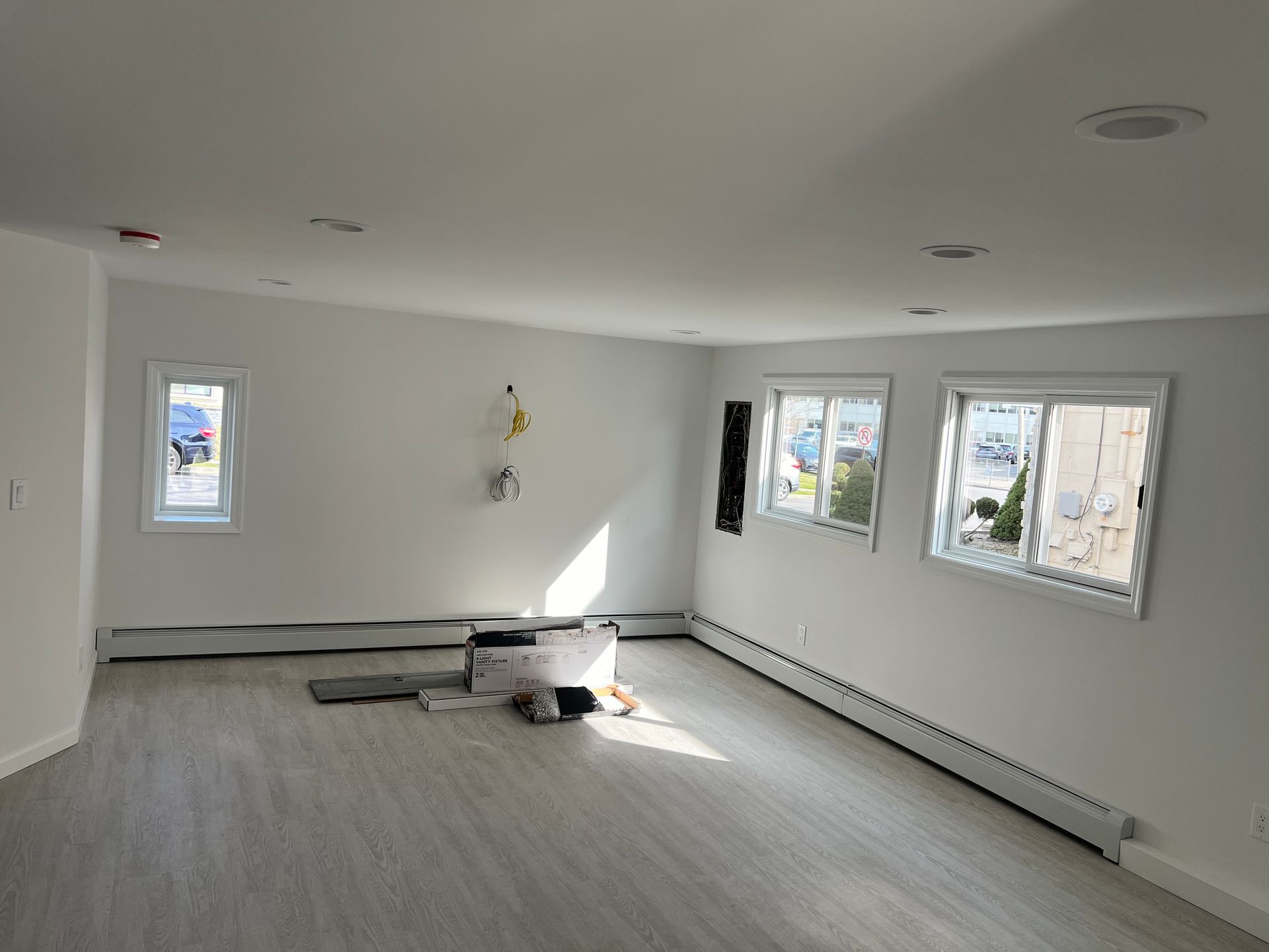 Empty room with white walls, three windows, and wood-look flooring. Radiators are along the bottom.