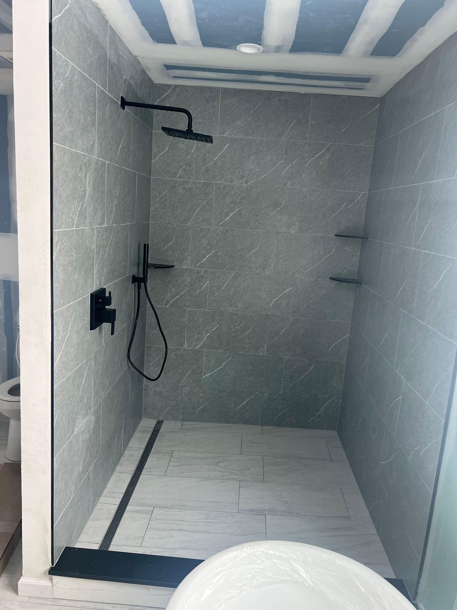 A gray tiled shower with black fixtures and shelving. The shower has a white floor with a black linear drain.