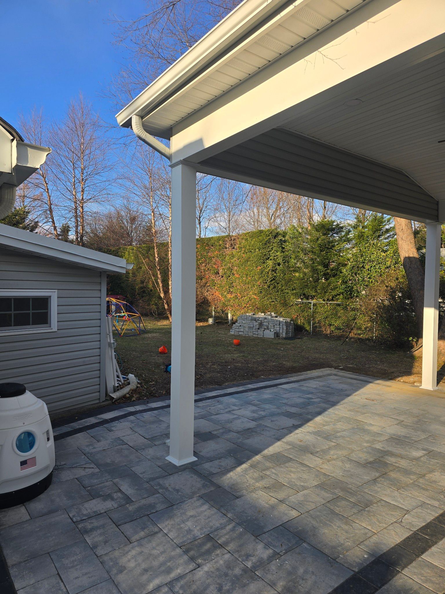 Gray patio with a white-columned awning overlooking a yard with a shed and trees.