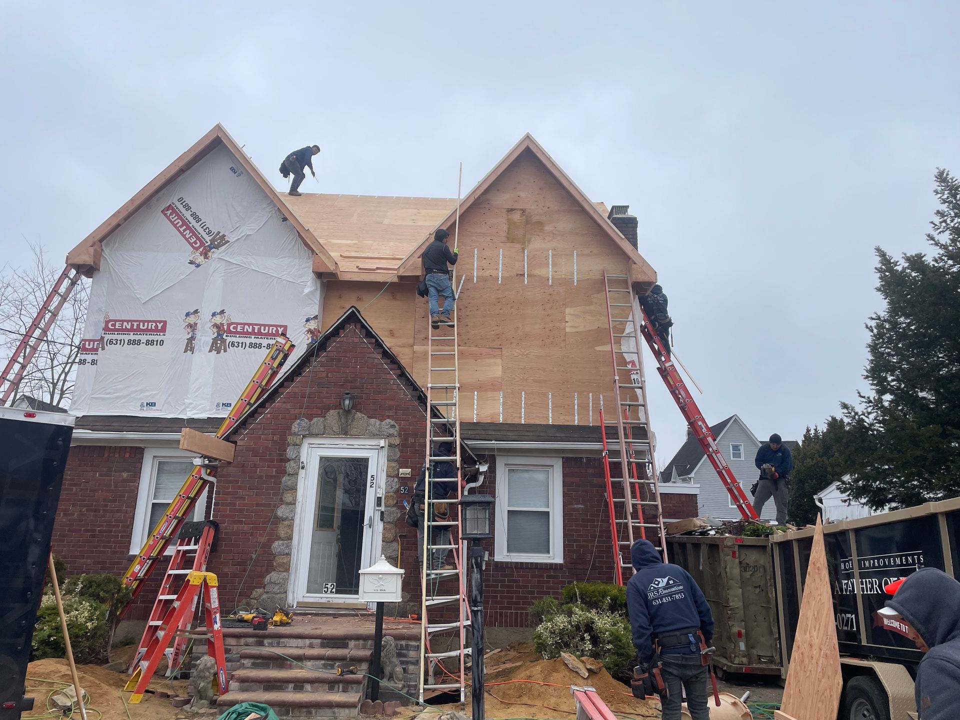 Construction workers on ladders renovating a two-story brick and wood-sided house; plywood, paper, and ladders are visible.