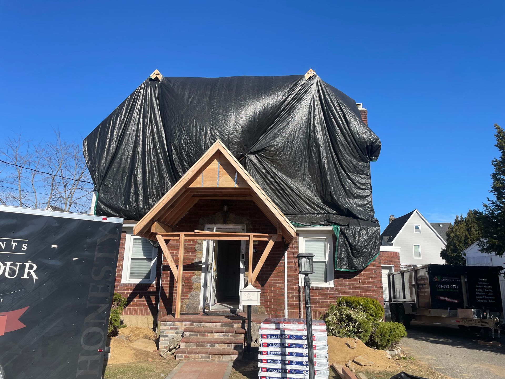 House with black tarp covering the roof during construction. Red brick, brown wooden porch, sunny blue sky.