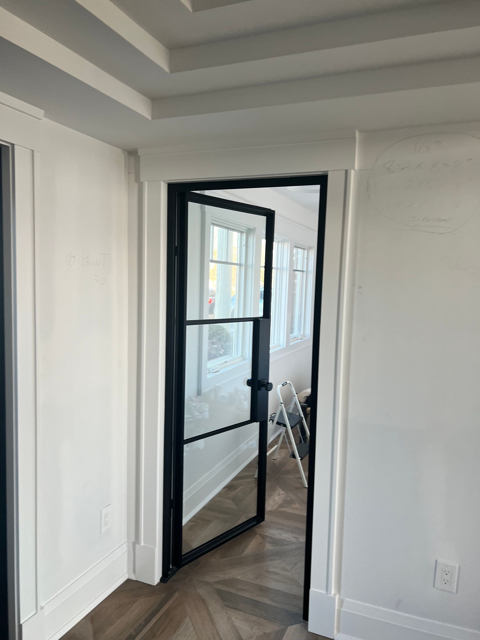 Black-framed glass door, open, in white-walled room with herringbone floor; sunlight through window visible.
