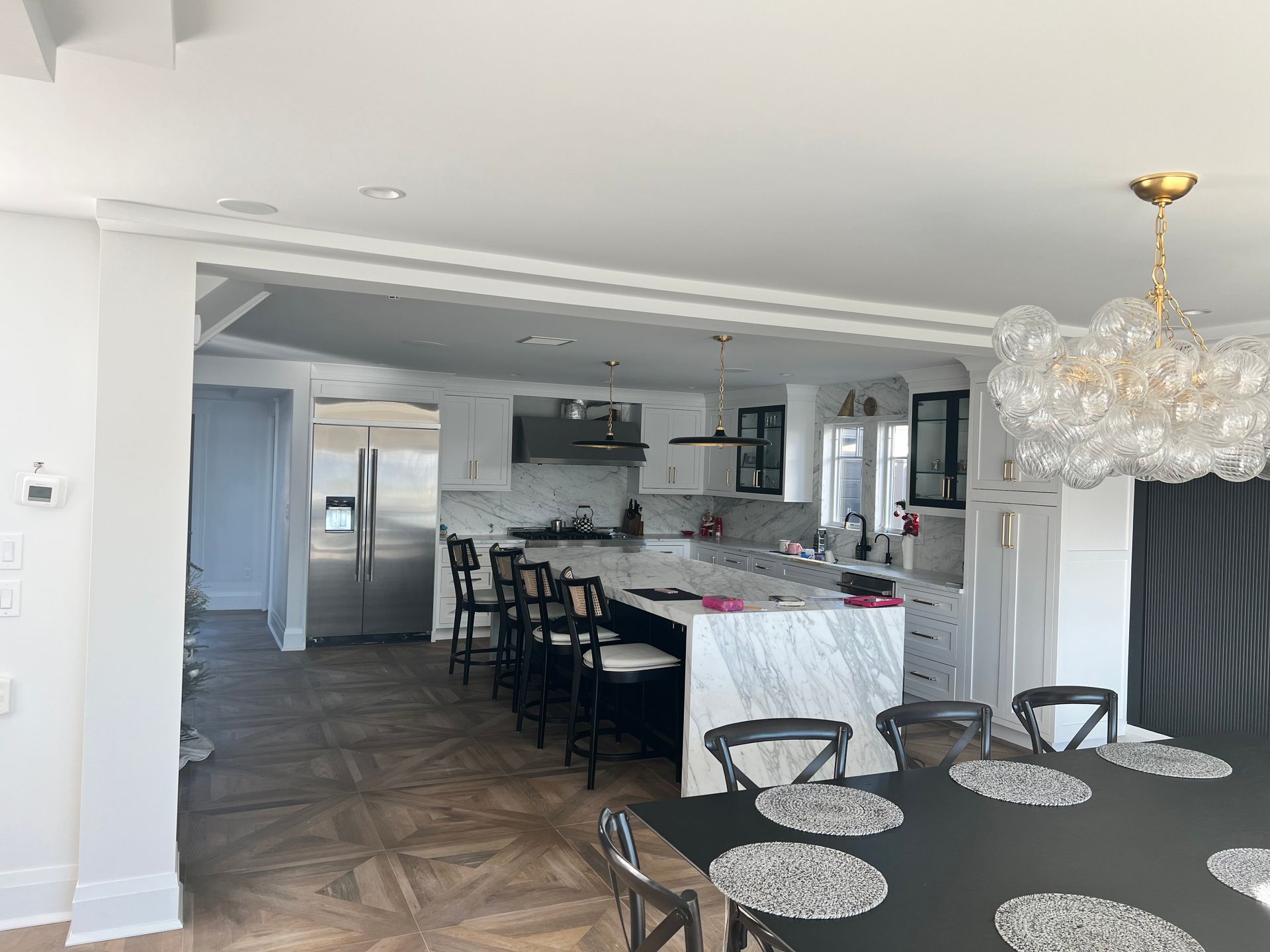 Modern kitchen with island and dining area. White cabinets, marble countertops, black stools, and gold chandelier.