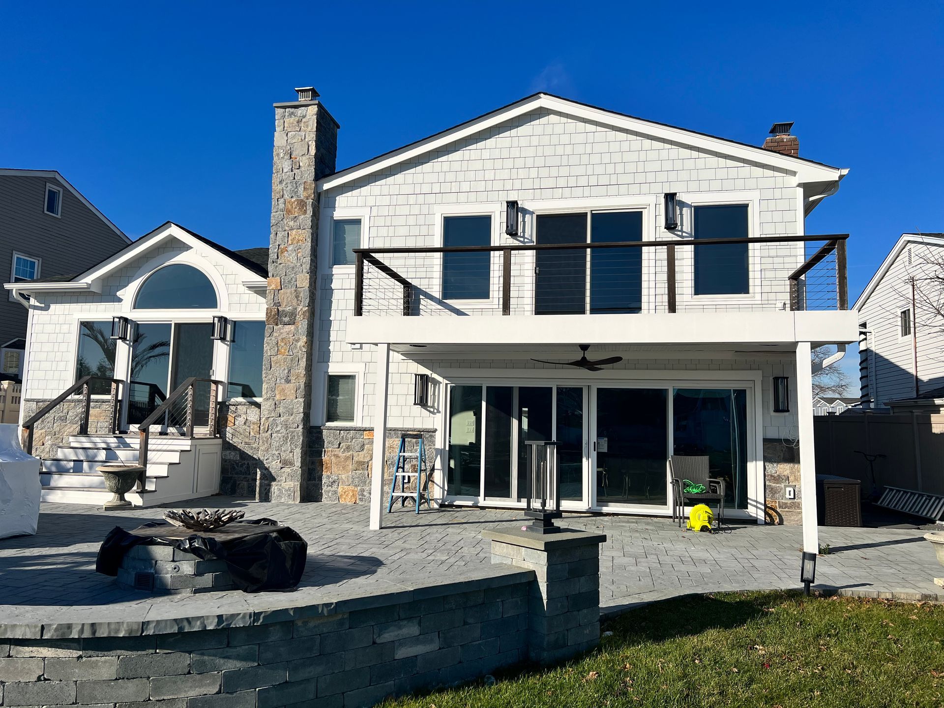 Two-story white house with a stone chimney and a backyard patio with a deck above, under a bright blue sky.
