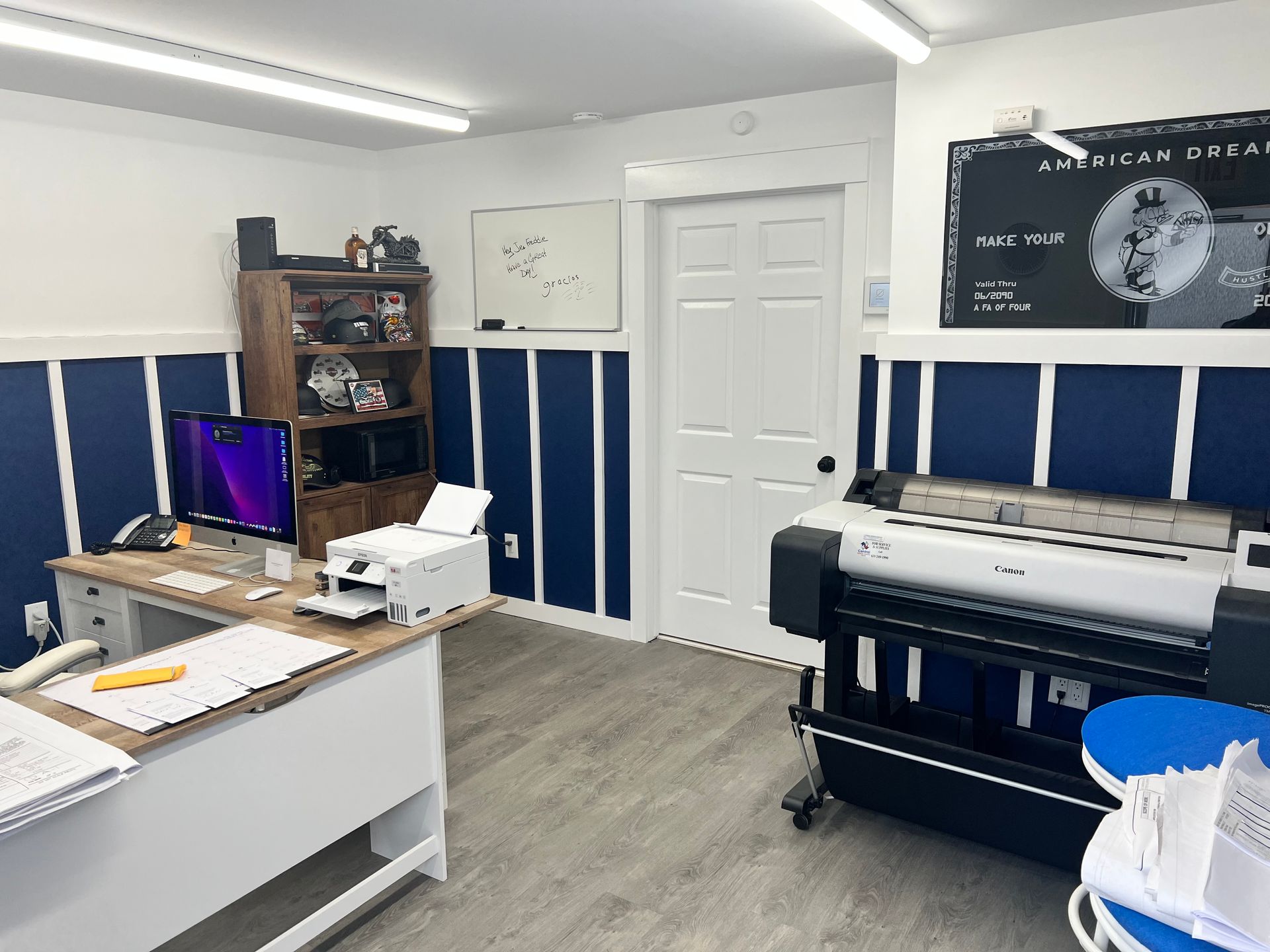Office with desk, printer, shelving unit, and large-format printer. Blue and white wall accents.