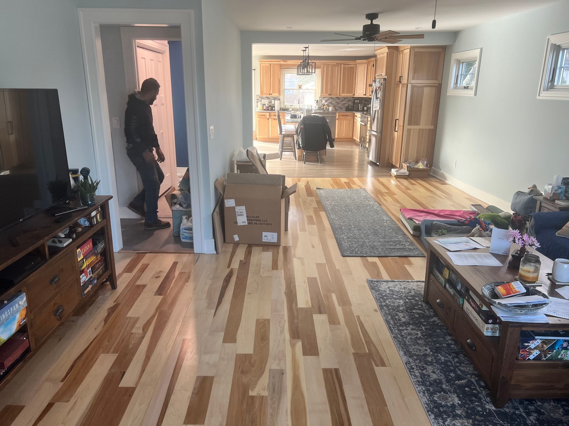Living room with hardwood floors, kitchen visible. A person walks through a doorway. Furnishings include a TV console, coffee table.