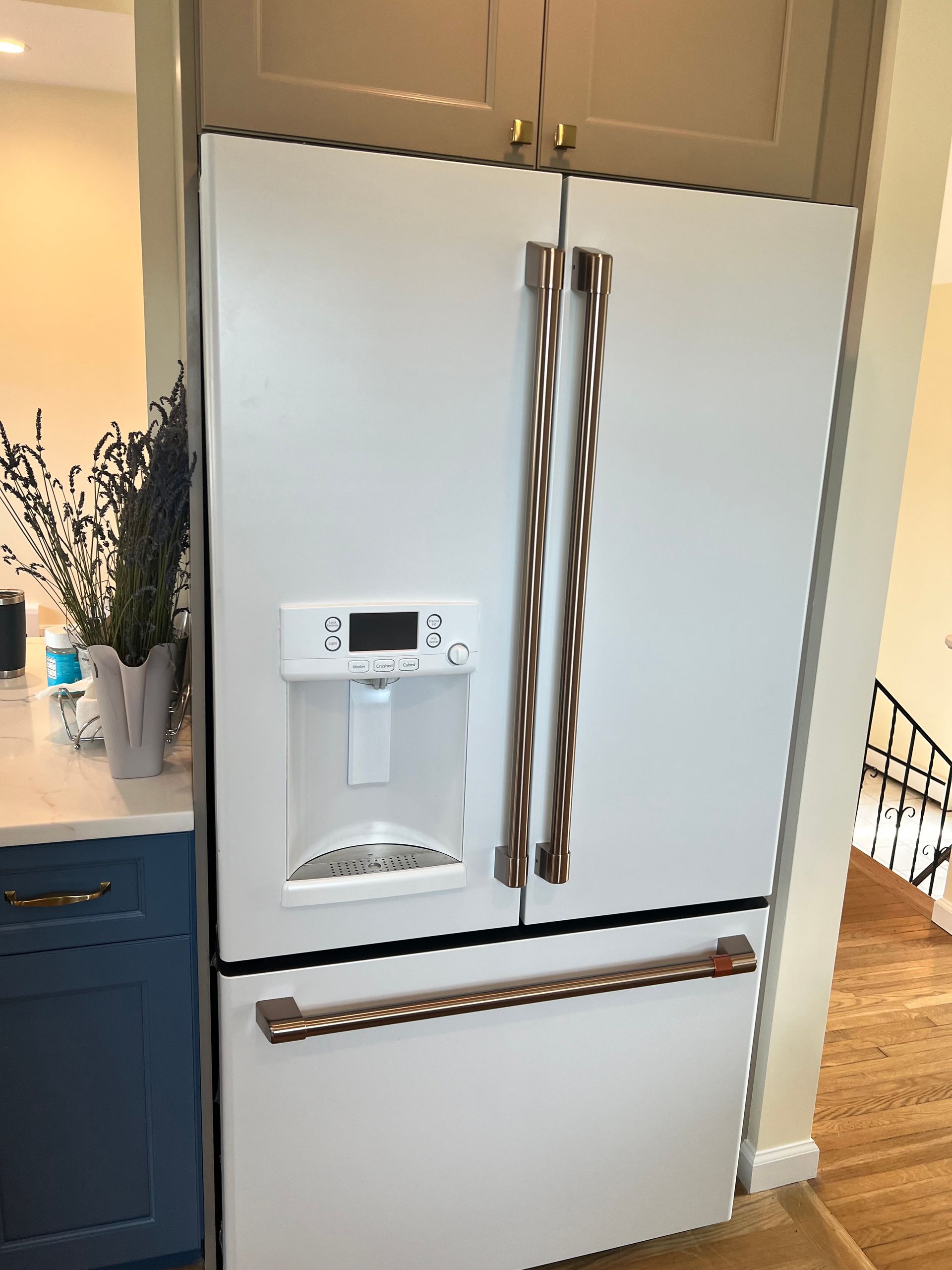 White French door refrigerator with bronze handles. Built-in, near a blue cabinet and stairs.