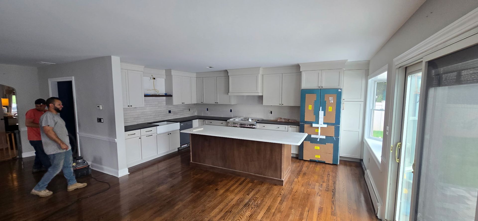 A partially renovated kitchen with white cabinets, dark countertops, a wood island, and two men standing near the entrance.