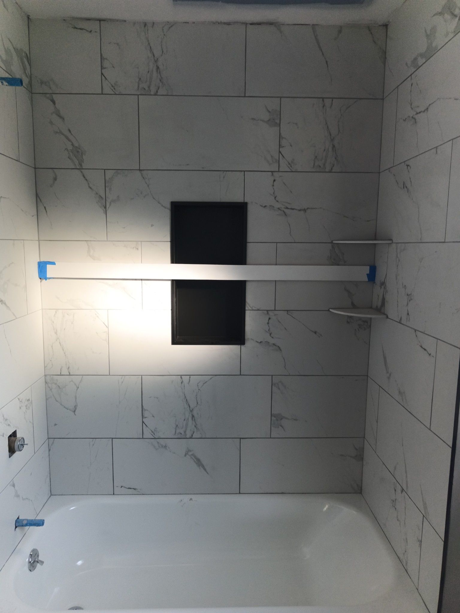 White tiled shower with a black shelf niche and silver bar, over a white bathtub.