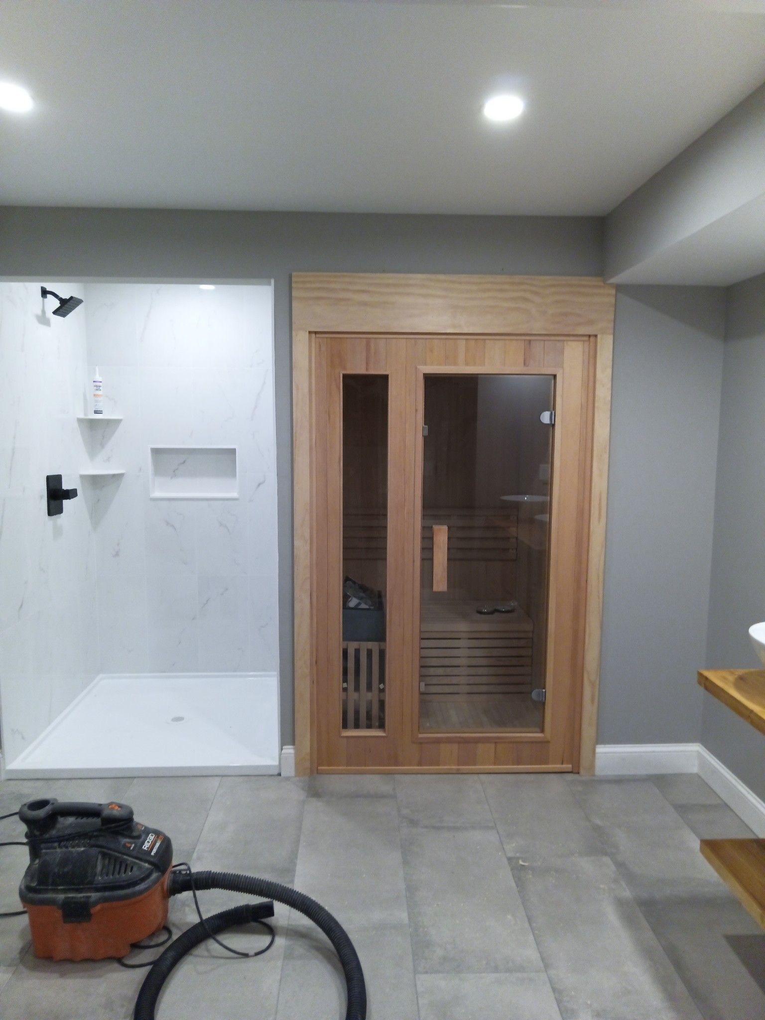 A newly constructed sauna and shower in a finished basement. The sauna has glass doors, and the shower is white.