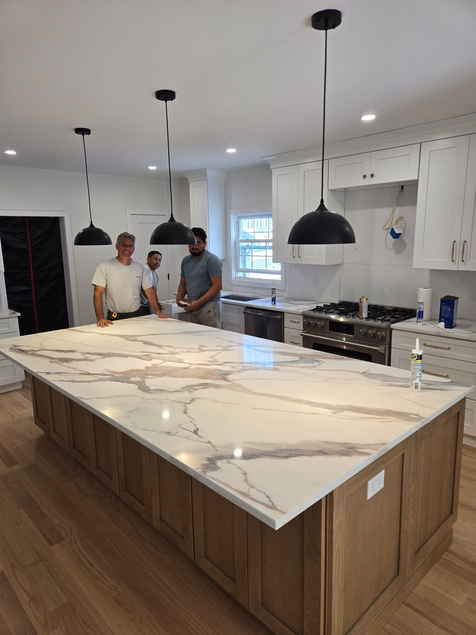 Three men in a new kitchen with an island. White marble countertop and black pendant lights. Wooden cabinets and flooring.