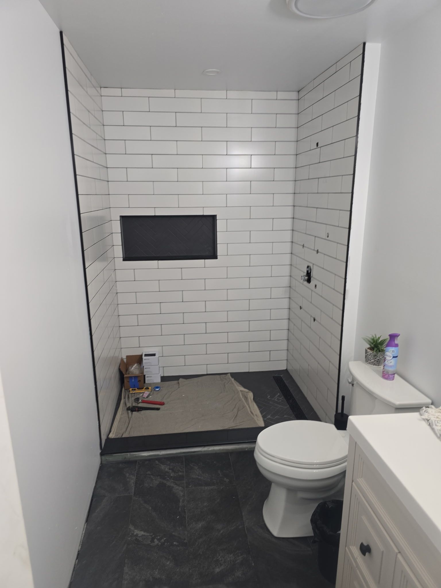 Bathroom under construction. White rectangular tiles in shower, dark floor. Toilet and vanity in room.