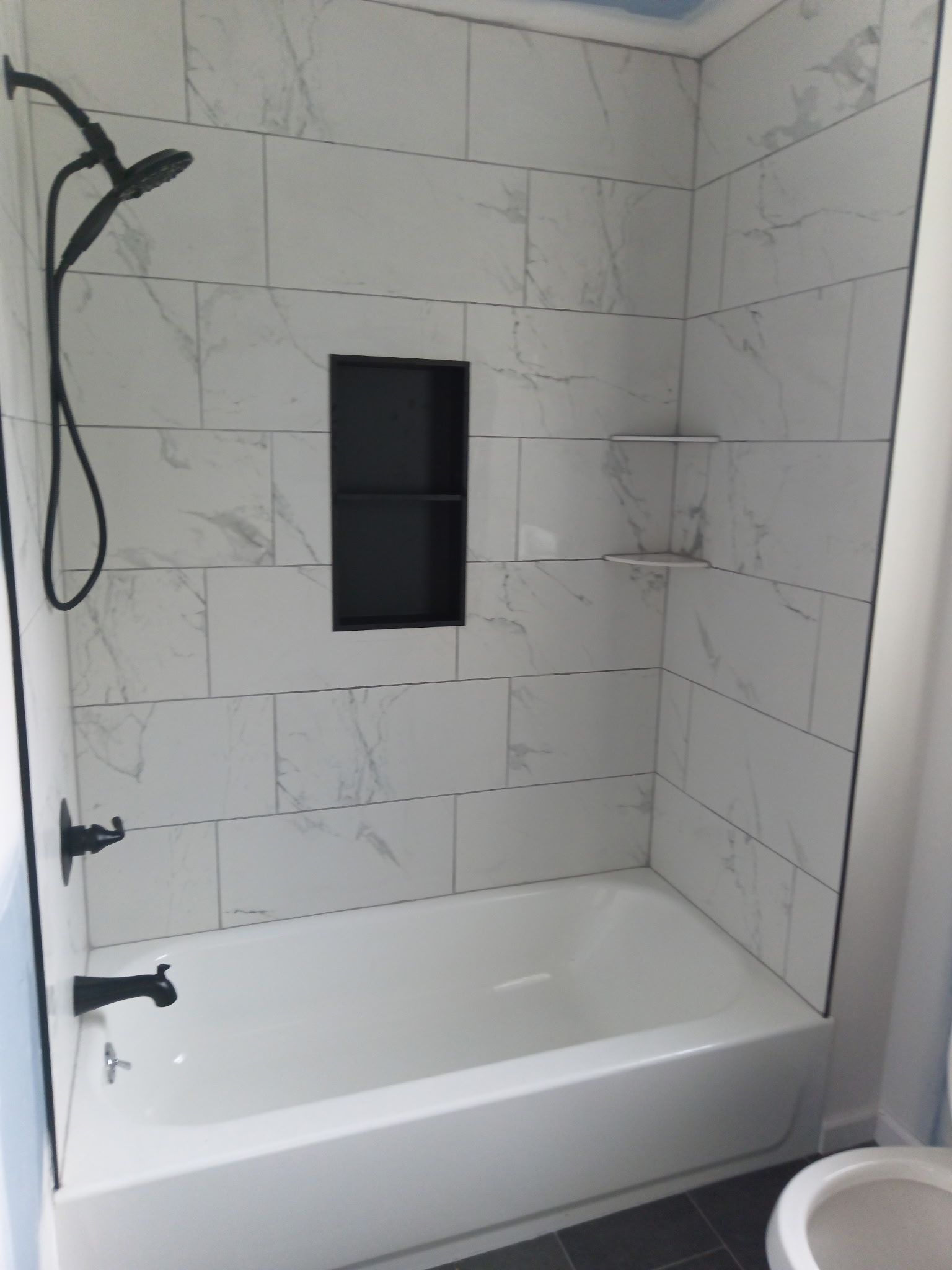 White tiled shower with black fixtures and built-in shelf and niche. Bathtub below.