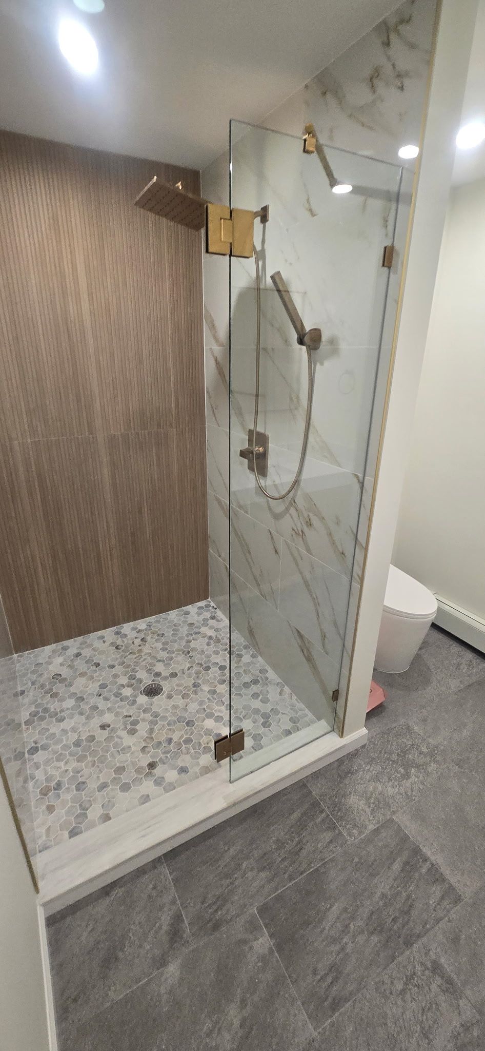 A modern shower with clear glass doors, gold fixtures, and textured stone floor.