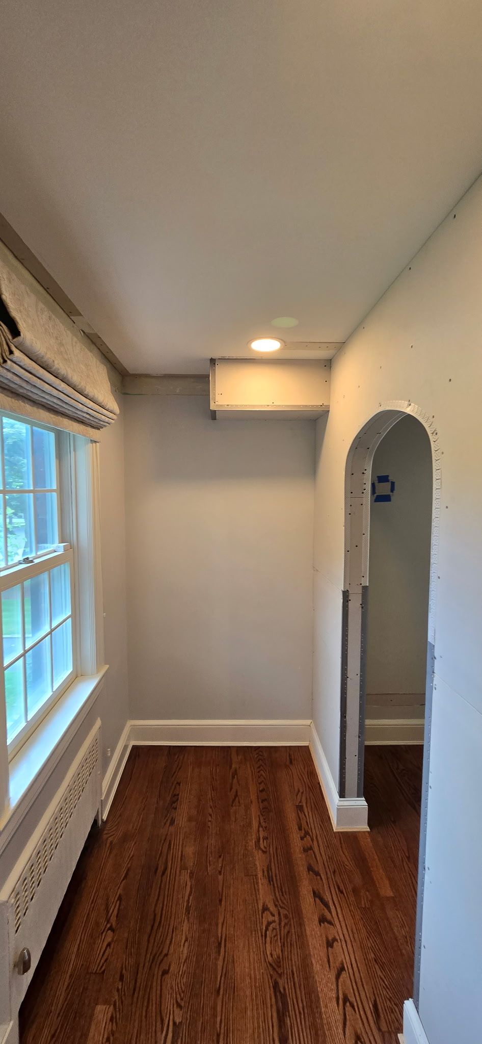 Narrow hallway with wood floors, window with blinds, and arched doorway.