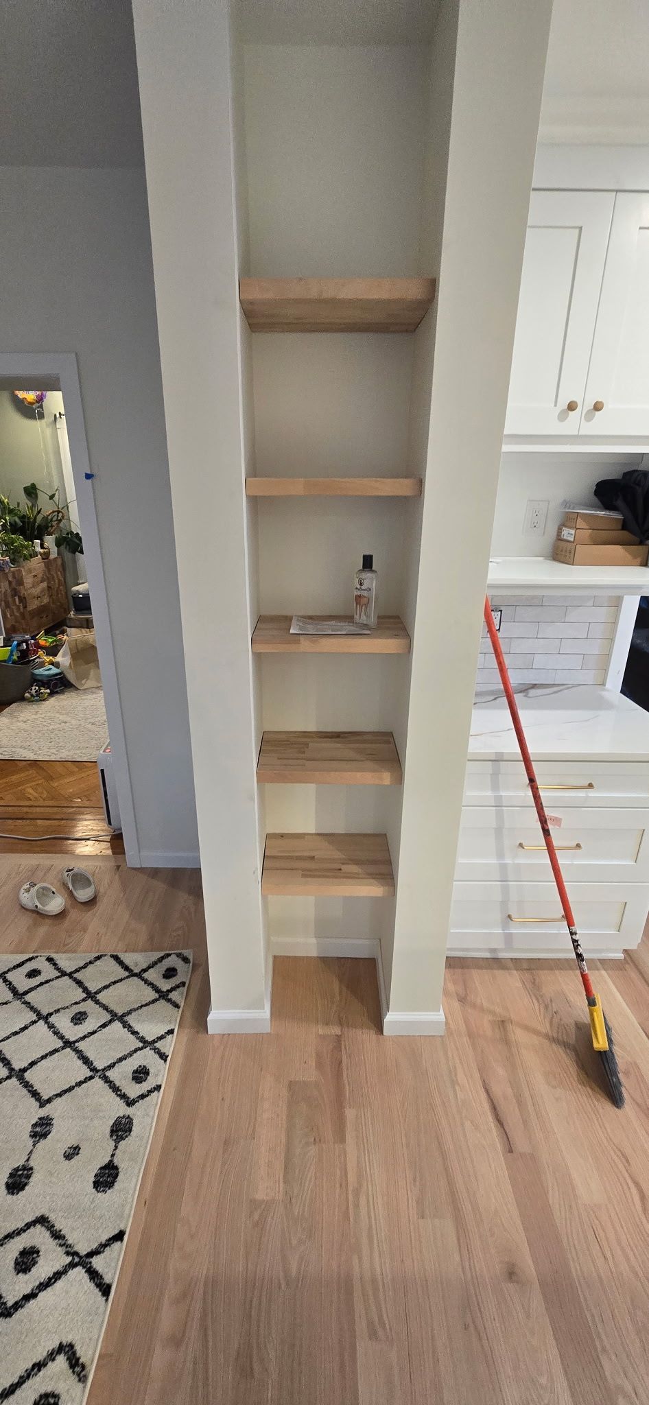 Built-in shelving unit with wooden shelves in a cream-colored wall, adjacent to a kitchen area.