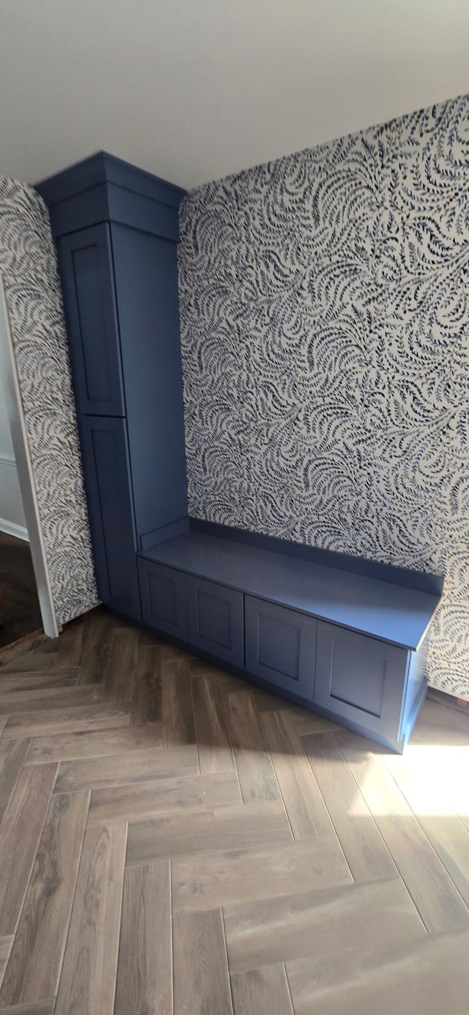 Blue built-in bench in corner of room with patterned wallpaper and herringbone floor.