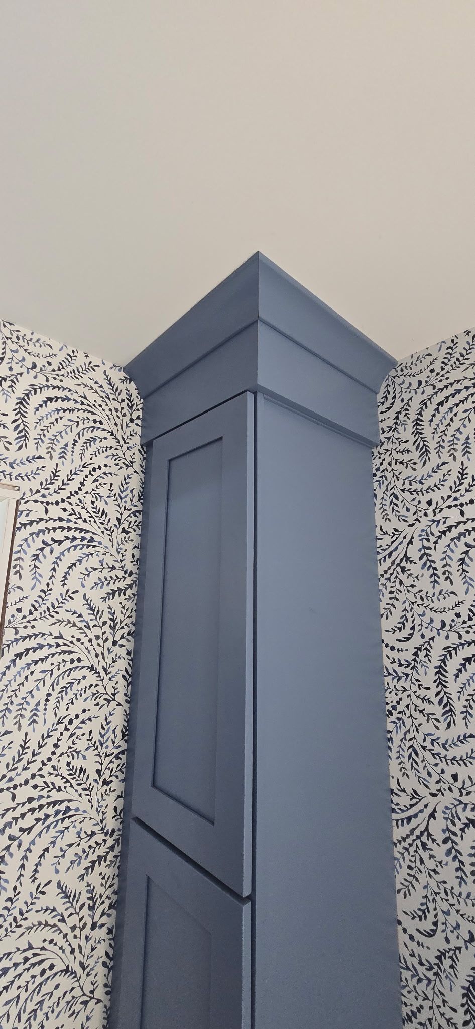 A tall, blue cabinet in a corner, with a decorative top, against a patterned wall and white ceiling.