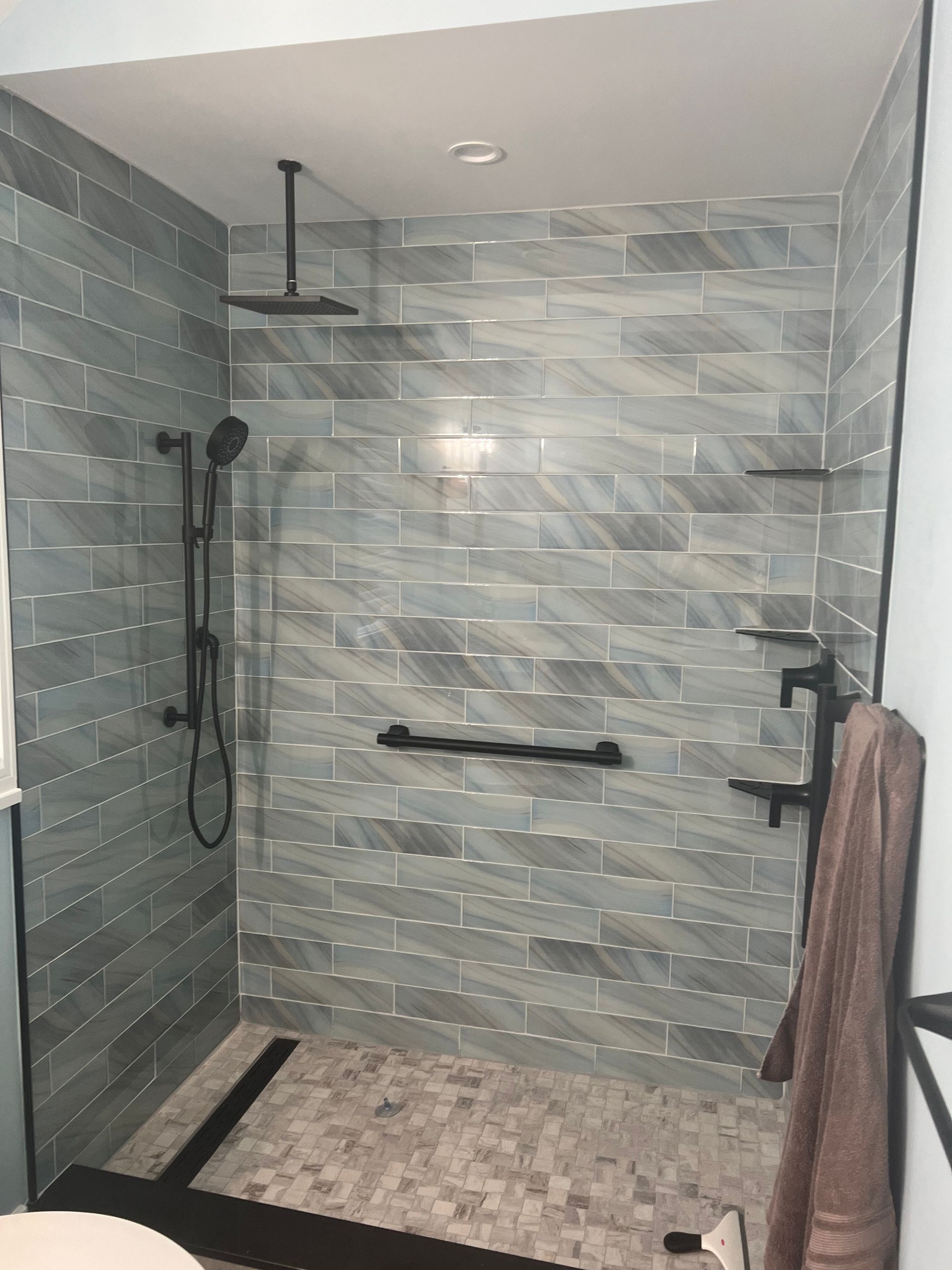 Modern shower with blue-grey tiled walls, black fixtures, and mosaic floor. A towel hangs on the right.