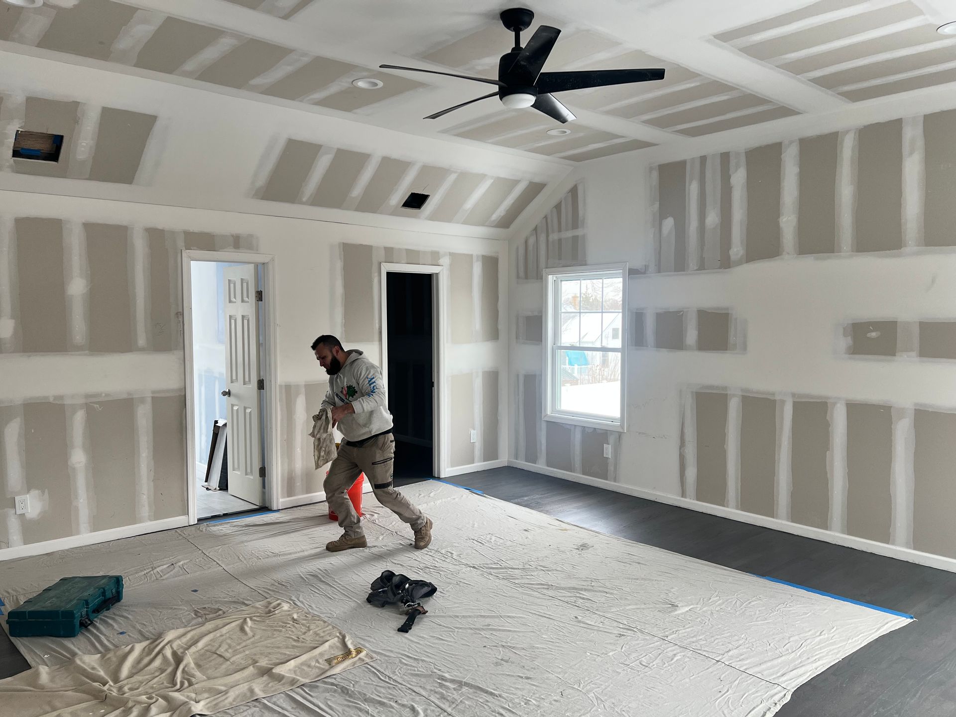 Room under construction; drywall installed. Person working, covered floor, black ceiling fan, and dark flooring.