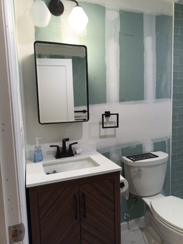 Bathroom renovation: vanity with black faucet and mirror, unfinished drywall, toilet, and tile.