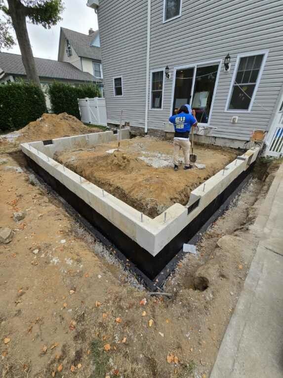 Construction of a rectangular foundation with a worker using a shovel. Grey house visible in the background.