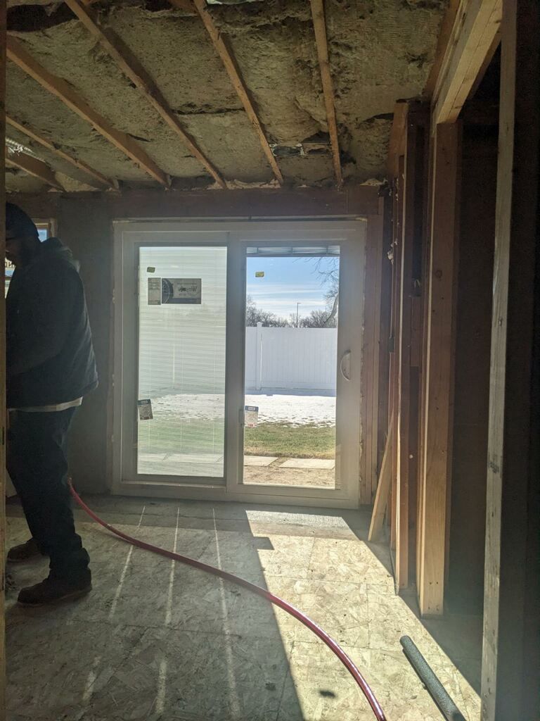 Interior of a room under construction with a sliding glass door. A person stands near the doorway, holding a red tube.