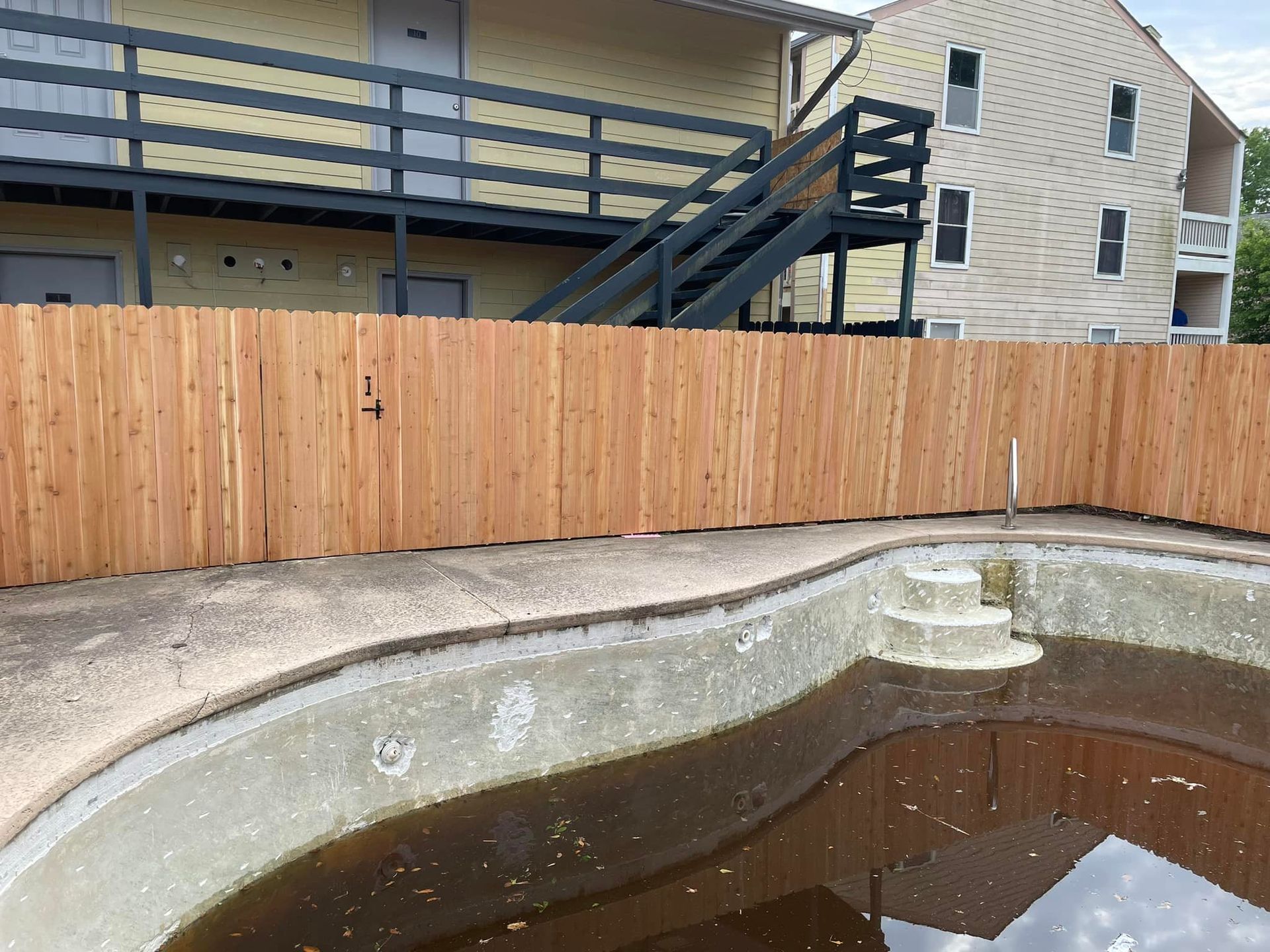 A wooden fence surrounds a murky, empty swimming pool near an apartment building.