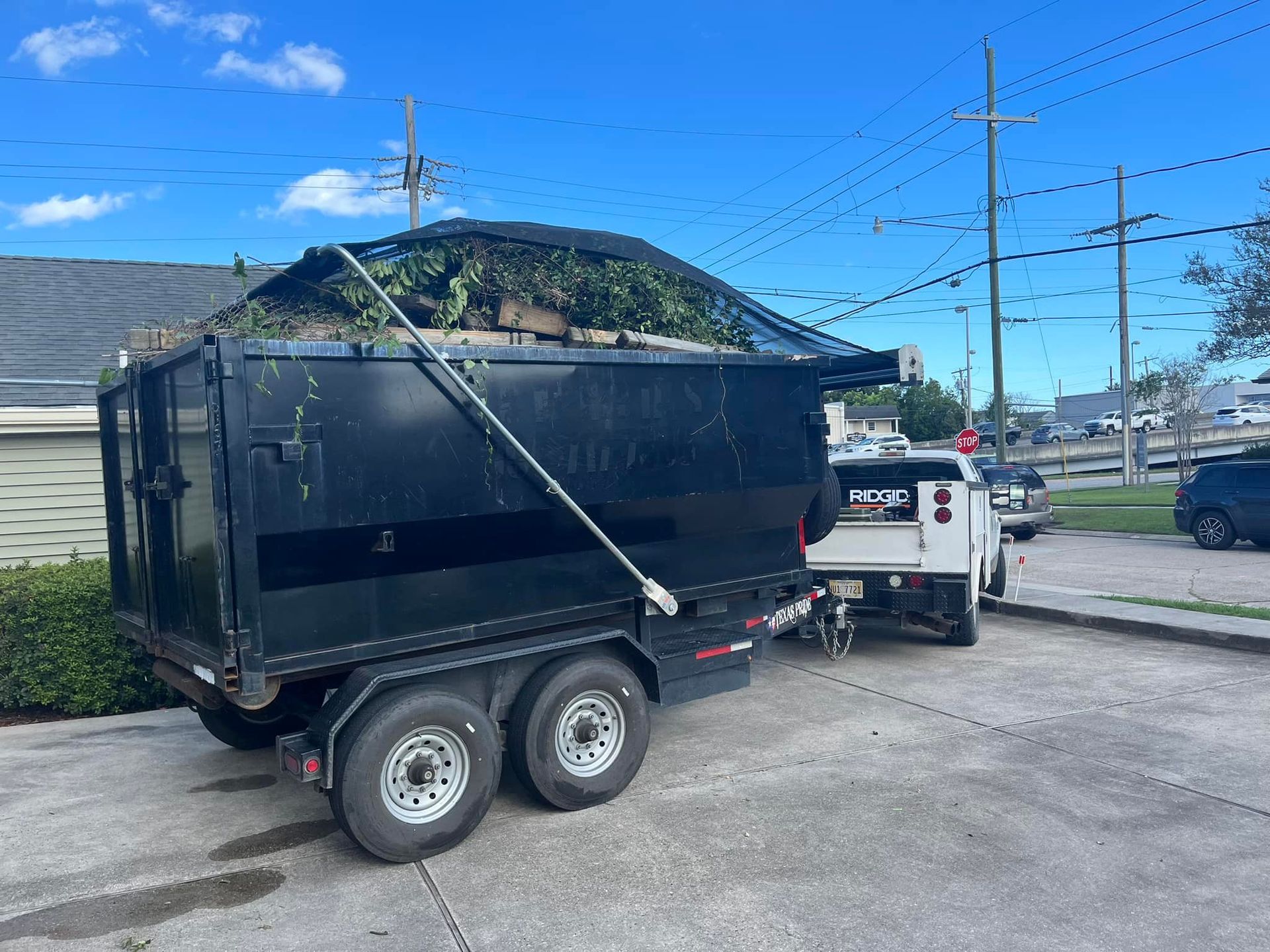 Black trailer filled with green debris, attached to a white truck, parked in a driveway under a blue sky.