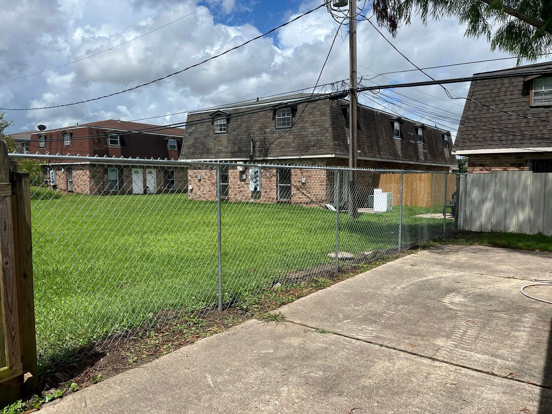 Chain link fence in front of a grassy yard, leading to brick buildings with dark roofs. Cloudy sky.
