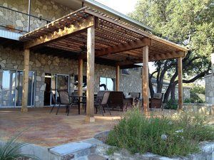 A wooden pergola is sitting on top of a patio next to a stone building.