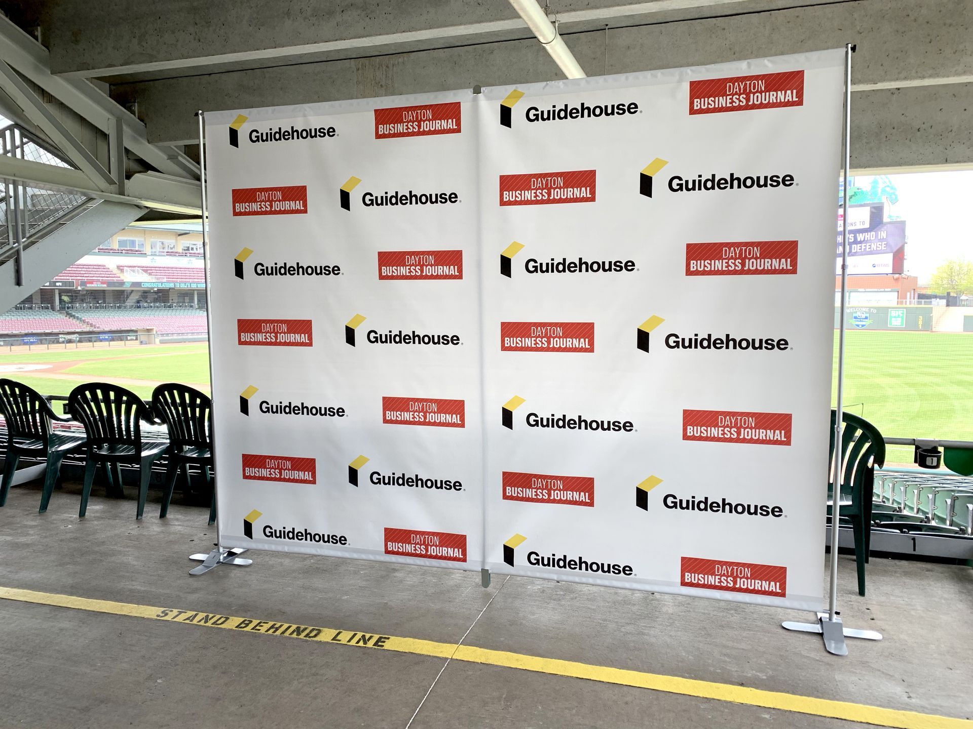 A two-panel Guidehouse backdrop at an outdoor event, with branding on a white background, near chairs and a stadium.