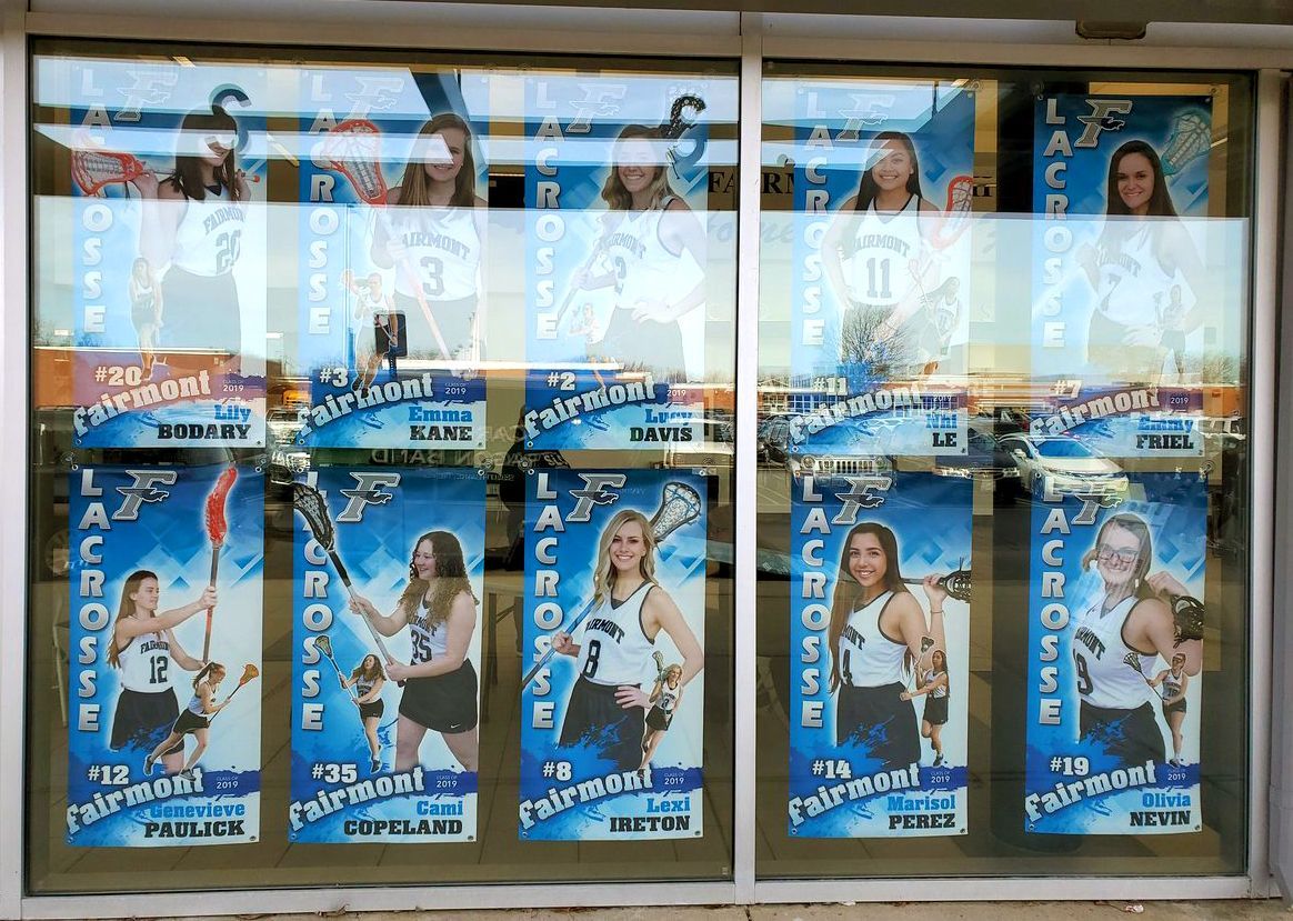 Window display of Fairmont lacrosse team posters featuring players in white uniforms with blue accents.
