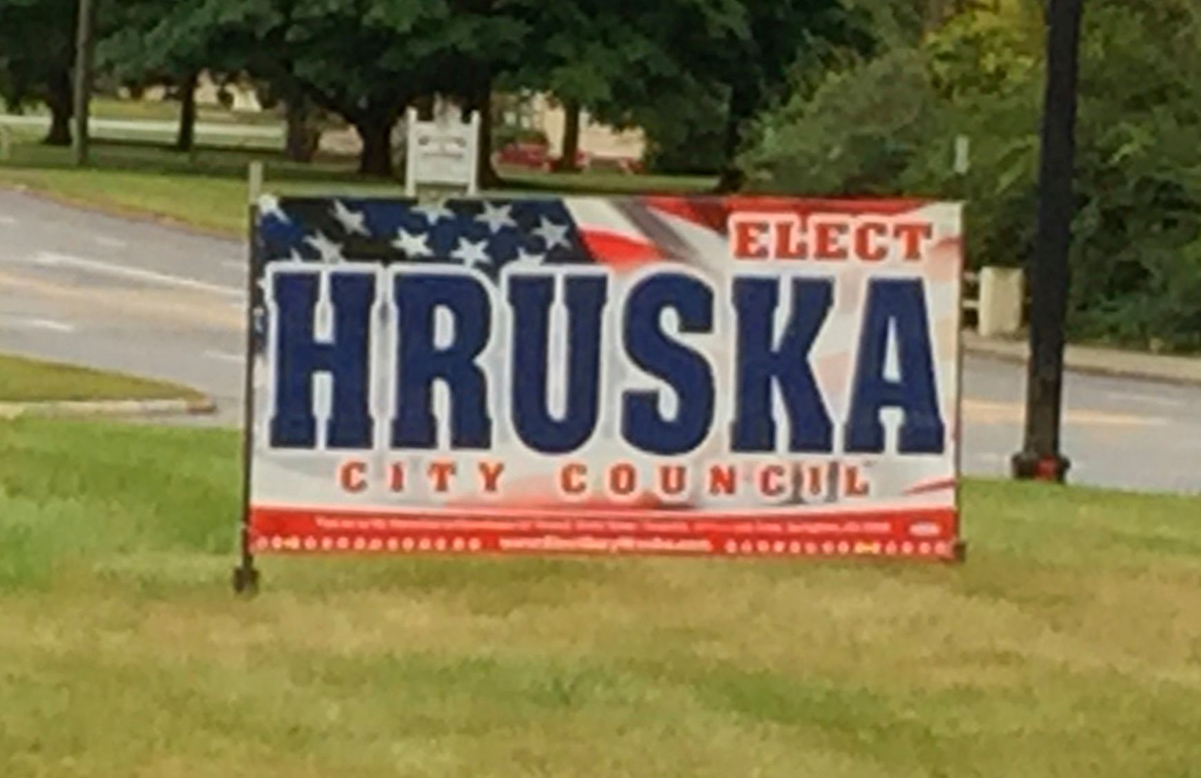 Sign for Hruska, candidate for City Council, with an American flag background.
