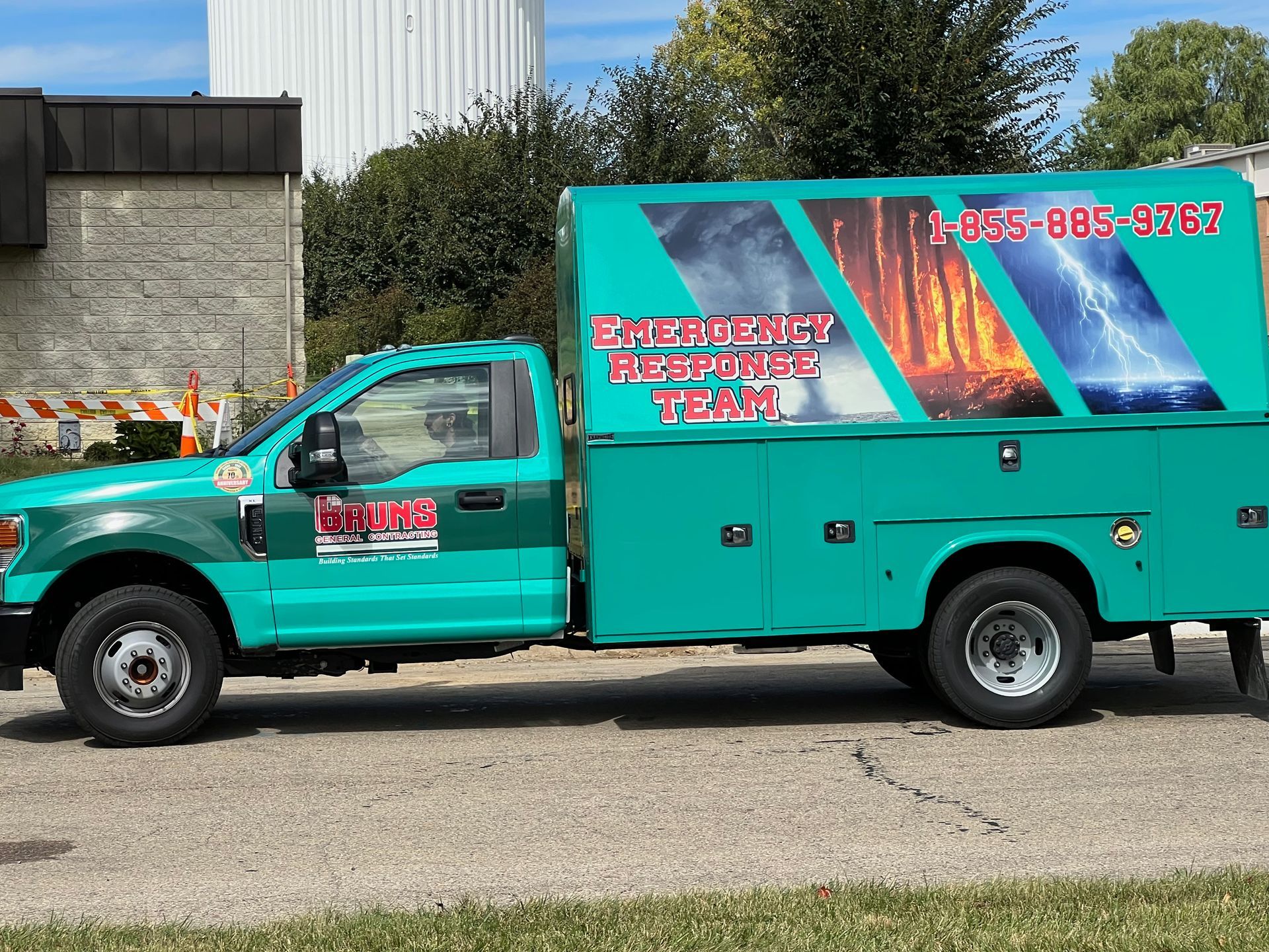 Teal work truck with emergency repair graphics, parked on asphalt, with a water tower in the background.