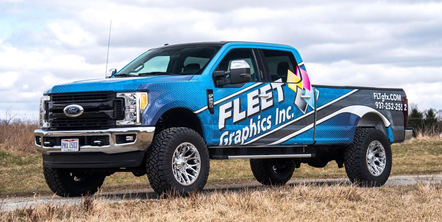 Blue Fleet Graphics Inc. truck, with custom graphics, parked on a grassy field, cloudy sky.