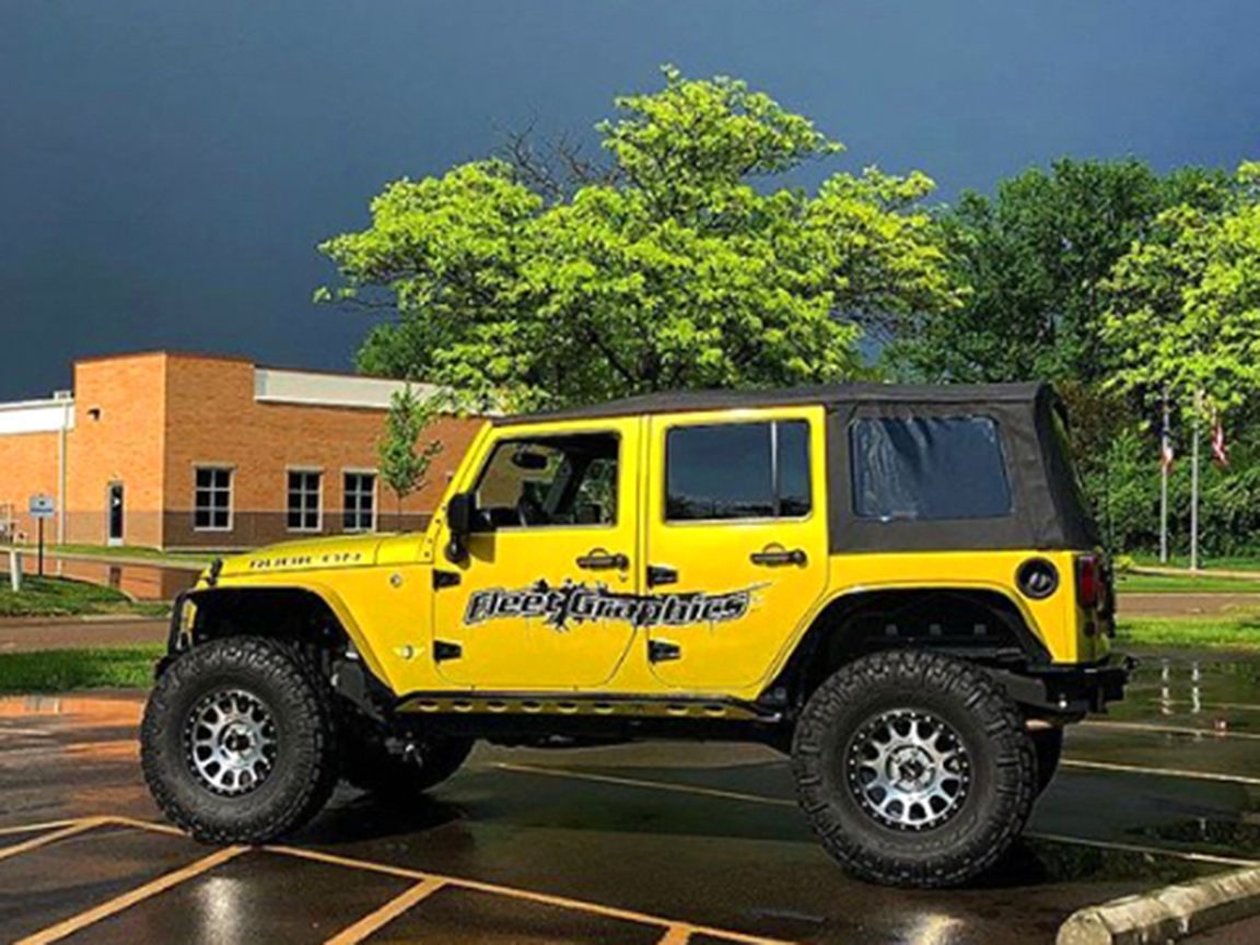 Yellow Jeep Wrangler, modified with large tires, parked in front of a building on a rainy day.