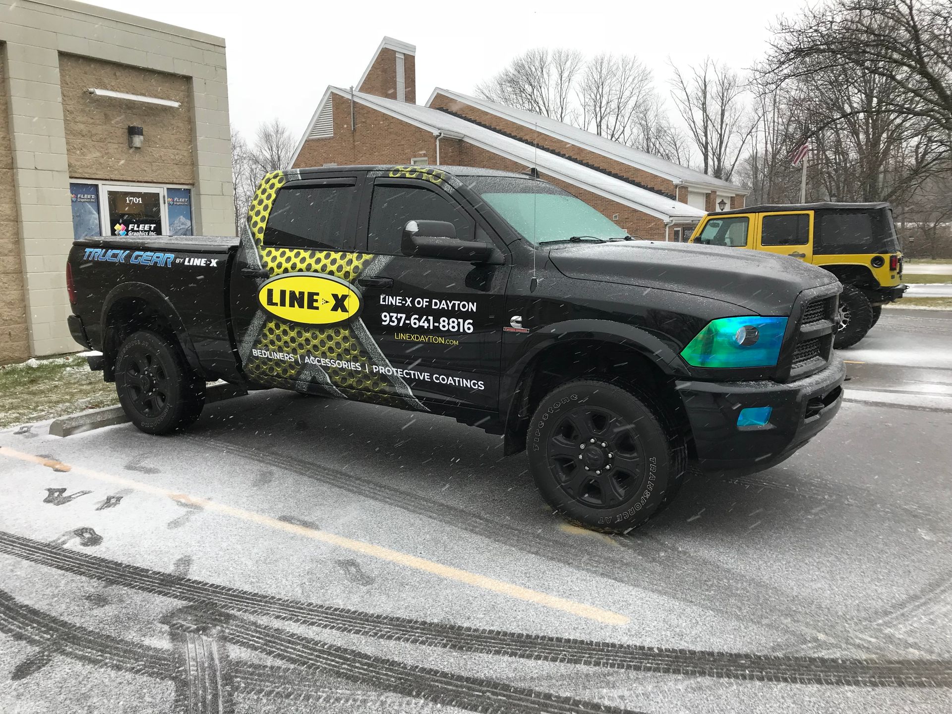 Black Line-X truck parked in snow, next to a yellow Jeep. Building in the background.