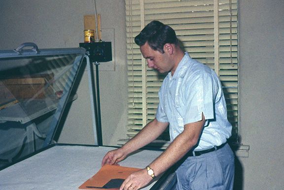 Man in light blue shirt and pants placing a dark square object on a large table with a glass cover. Window with blinds in background.