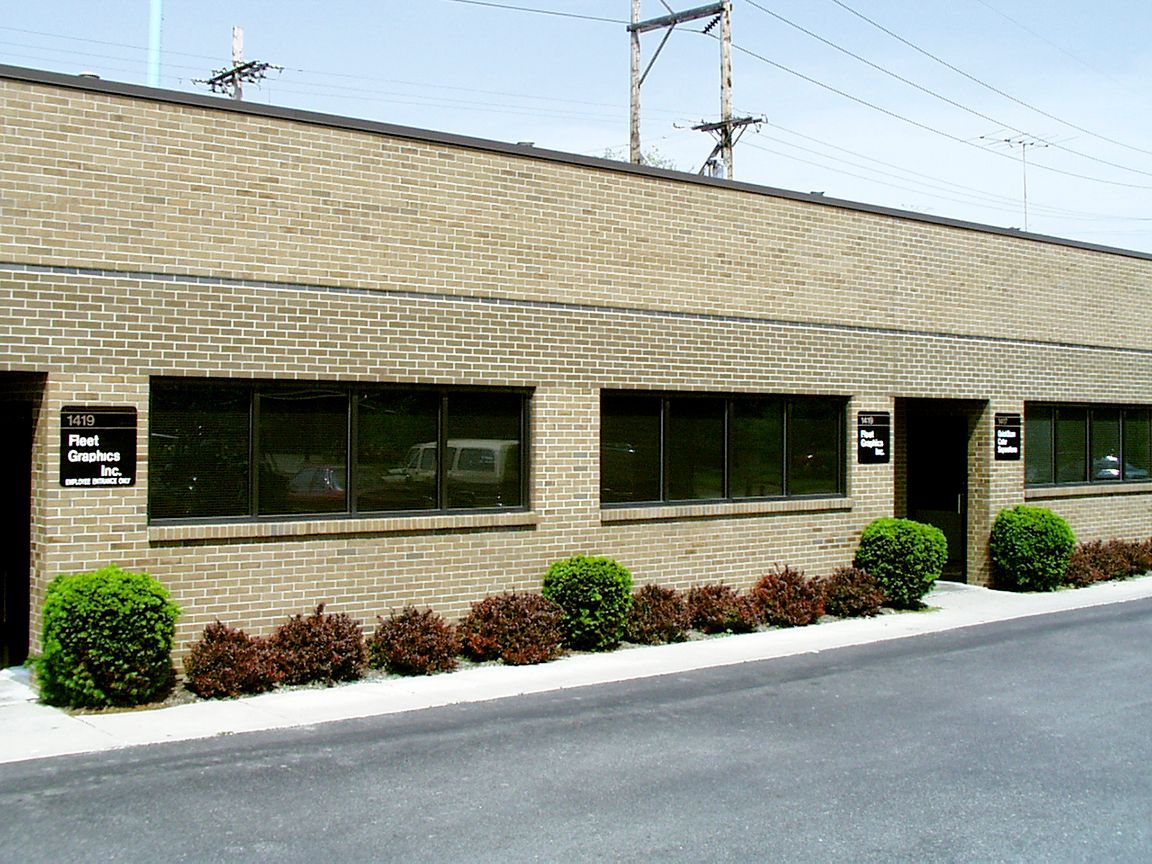 Tan brick building with dark windows, green bushes, and a paved driveway.