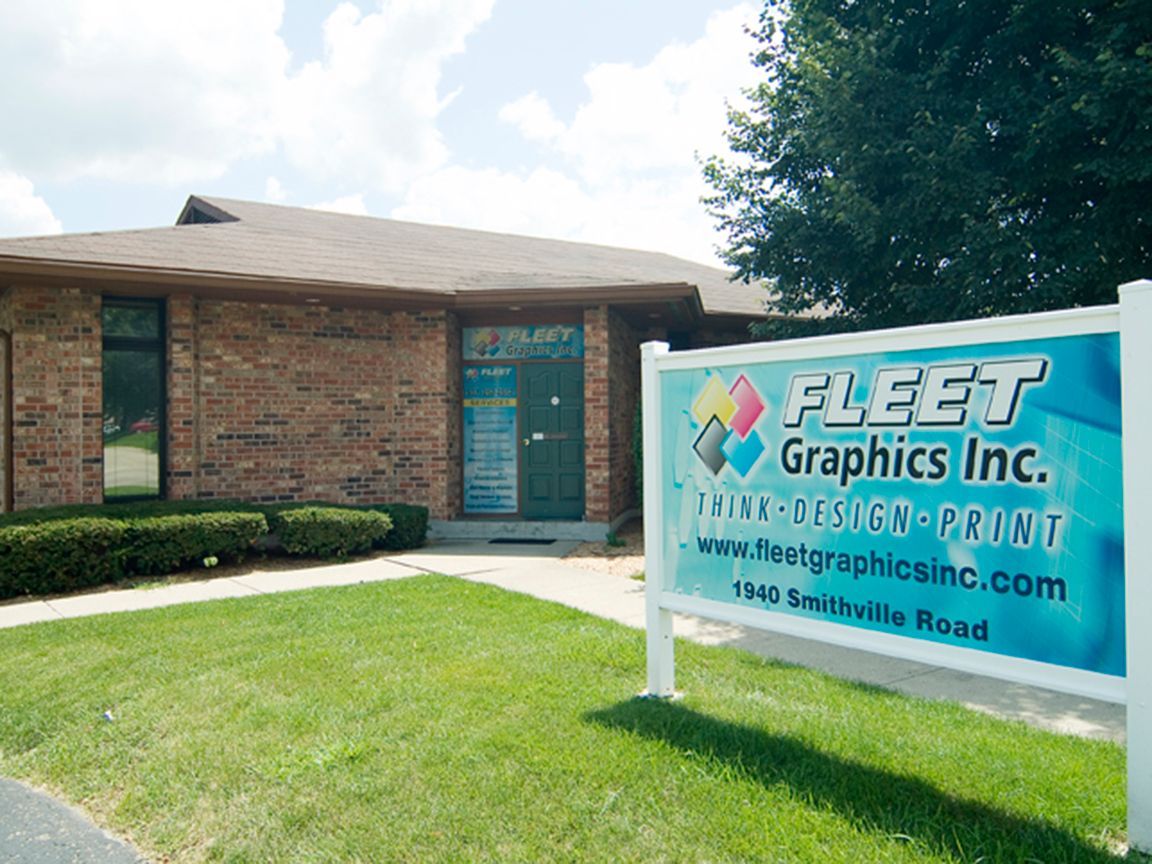 Fleet Graphics Inc. sign in front of a brick building on a sunny day; green grass, bushes, and a website address.